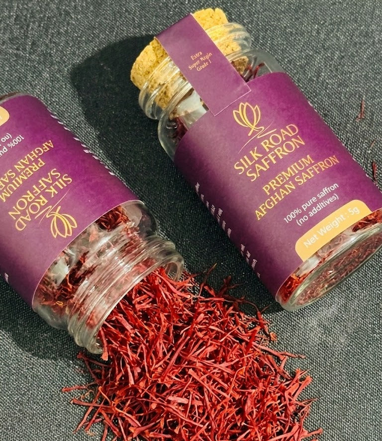 Two Silk Road Saffron 5g glass jars with deep red Afghan saffron threads, one jar open with threads spilling onto a dark textured surface.