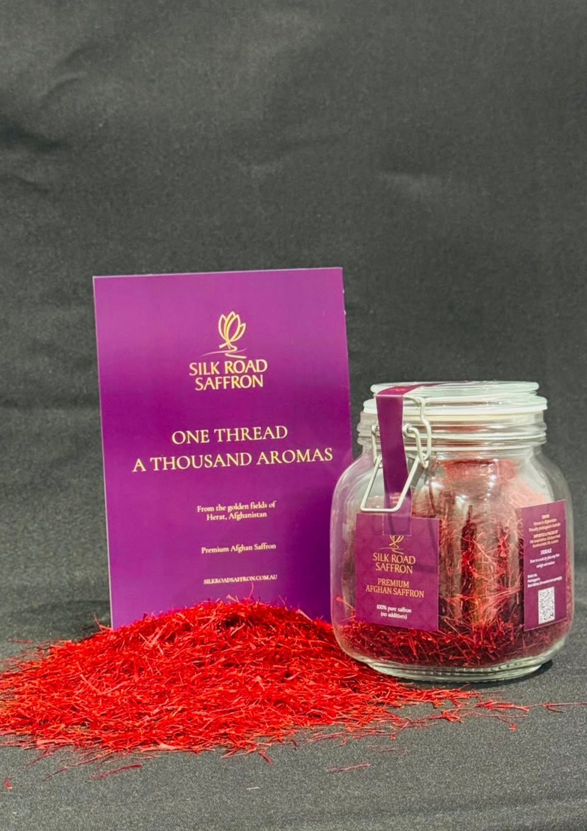 Silk Road Saffron 'One Thread A Thousand Aromas' product display featuring a purple brand card, a glass jar filled with red saffron threads, and a pile of loose red saffron on a dark gray background.