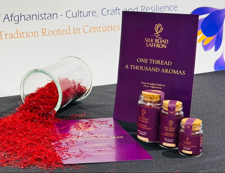 Silk Road Saffron products displayed on a dark tabletop, including large and small glass jars filled with deep red saffron threads, a spilled pile of red saffron, a purple product box, and three small saffron bottles. A white backdrop with 'SILK ROAD SAFFRON' text is visible behind the items.