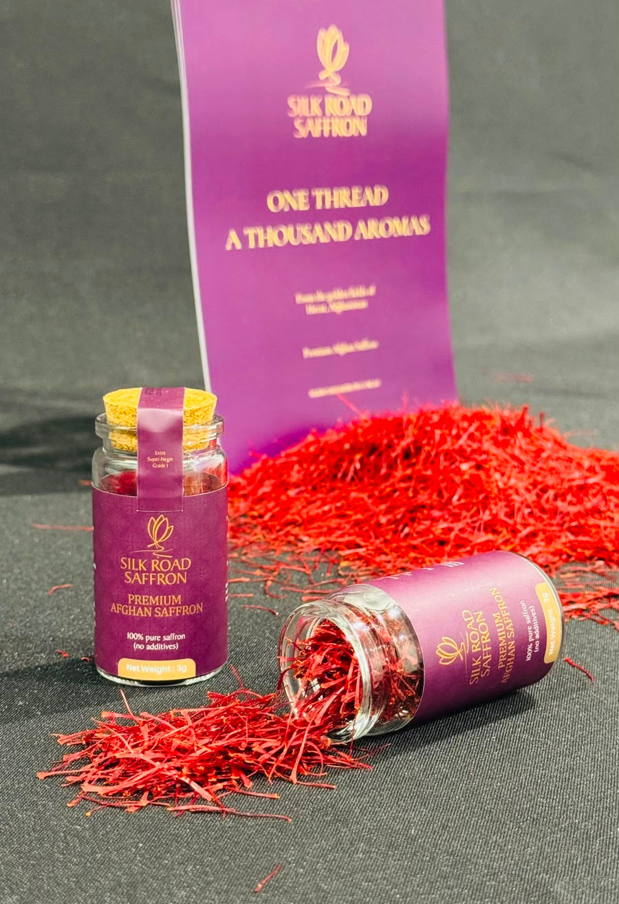 Two small jars of Silk Road Saffron with purple labels are displayed on a dark gray surface, with loose red saffron strands scattered around them and a tall purple product card in the background.