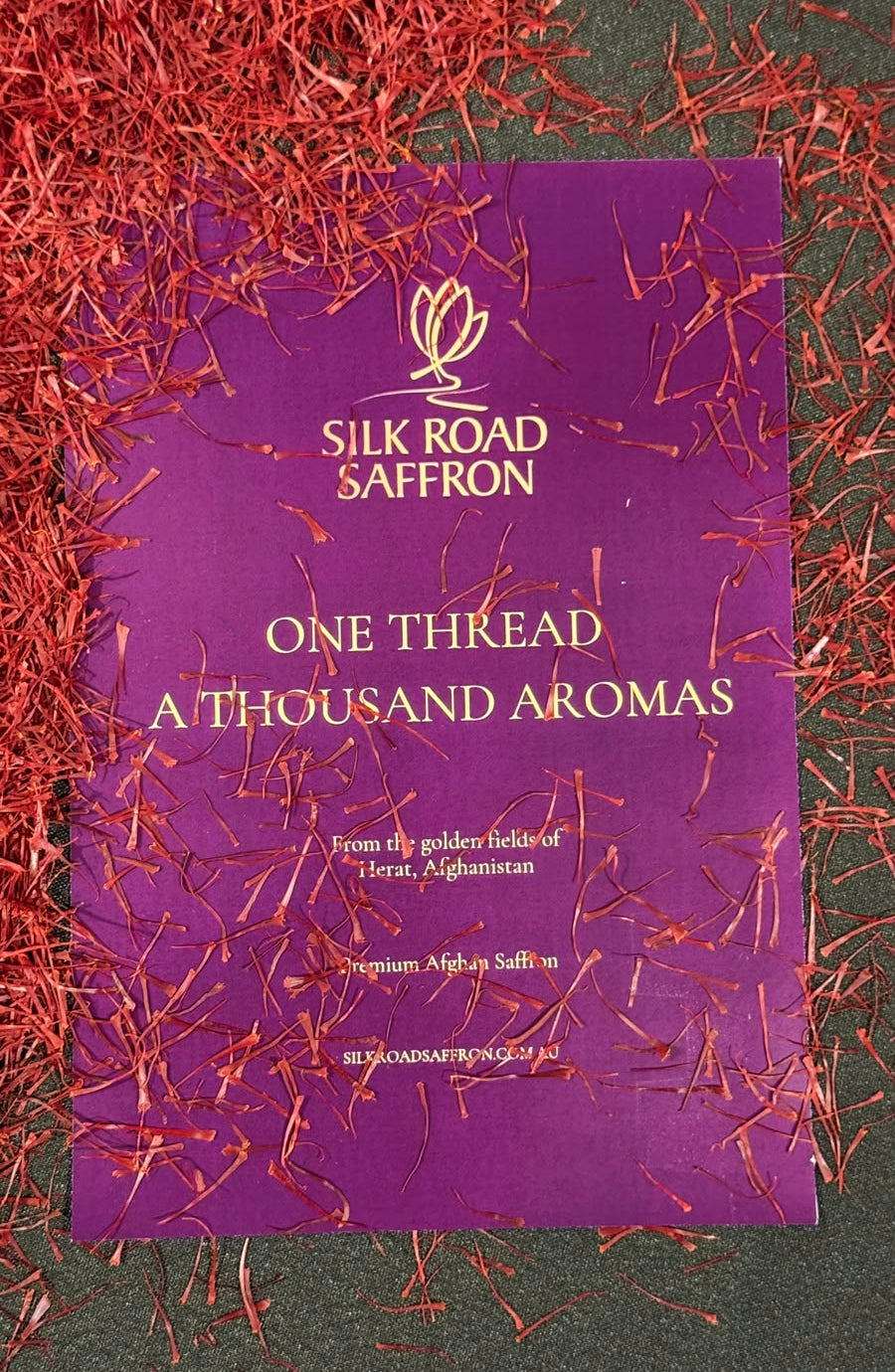 A purple brand paper titled 'Silk Road & Saffron One Thread: A Thousand Aromas' lies on a dark green textured surface surrounded by shredded red saffron threads.