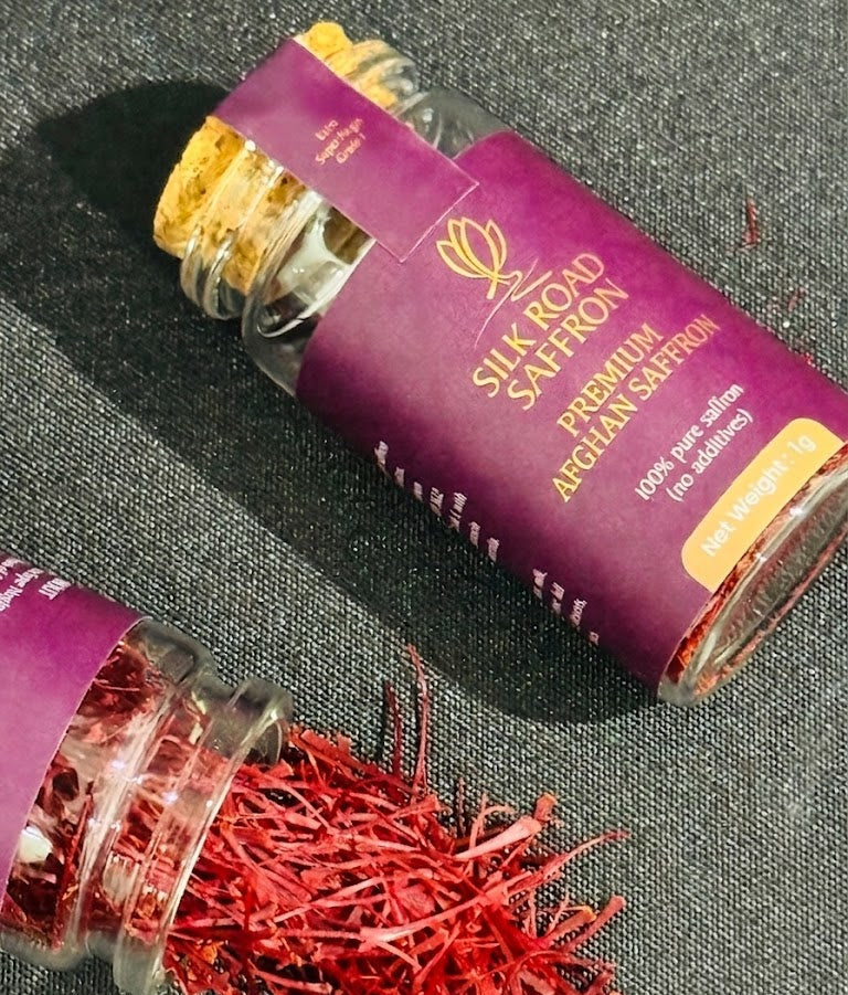 Two clear glass jars of Silk Road Saffron with purple labels, one lying on its side with red saffron threads spilling out onto a dark gray textured surface.