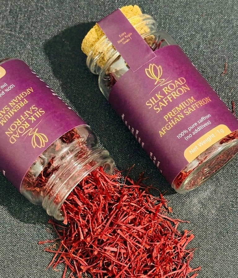 Two Silk Road Saffron 1g glass jars with deep red Afghan saffron threads, one jar open with threads spilling onto a dark textured surface.