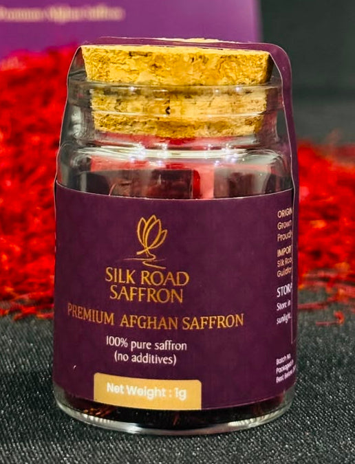 A small glass jar of Silk Road Saffron with a gold foil lid and purple label, placed in front of a larger purple branded box, with scattered red saffron threads on a dark surface.