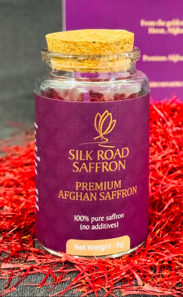 A glass jar of Silk Road Saffron with a cork stopper and a purple label, placed in front of a matching purple product box. The jar is surrounded by loose red saffron threads on a dark textured surface.