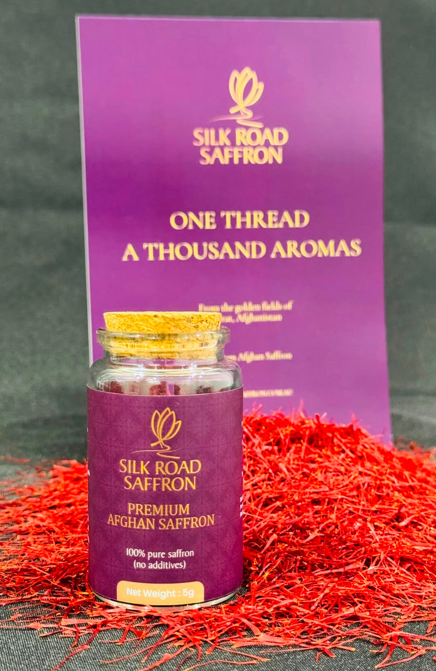 A 5 gram saffron jar of Silk Road Saffron with a purple label and gold cap, placed in front of a matching purple product card. Red saffron threads are scattered around the jar on a dark textured background.