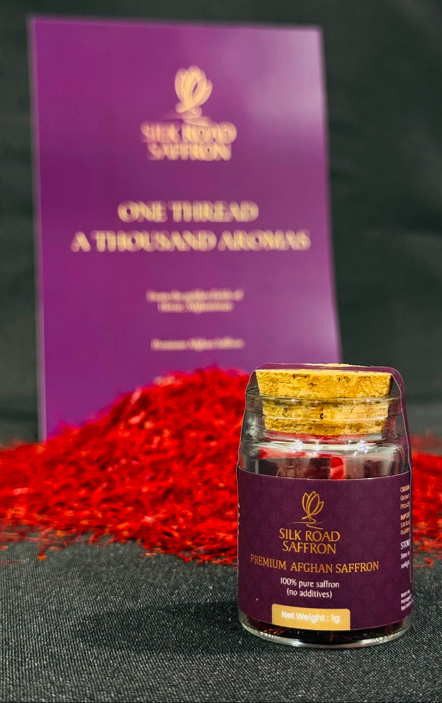 A small glass bottle of Silk Road Saffron with a gold cap and dark purple label, placed in front of a larger purple branded card, with red saffron threads scattered on a dark surface.