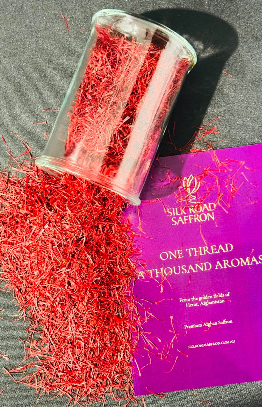 Red saffron threads spilled from a clear glass jar onto a dark gray surface, next to a purple Silk Road Saffron brand card.